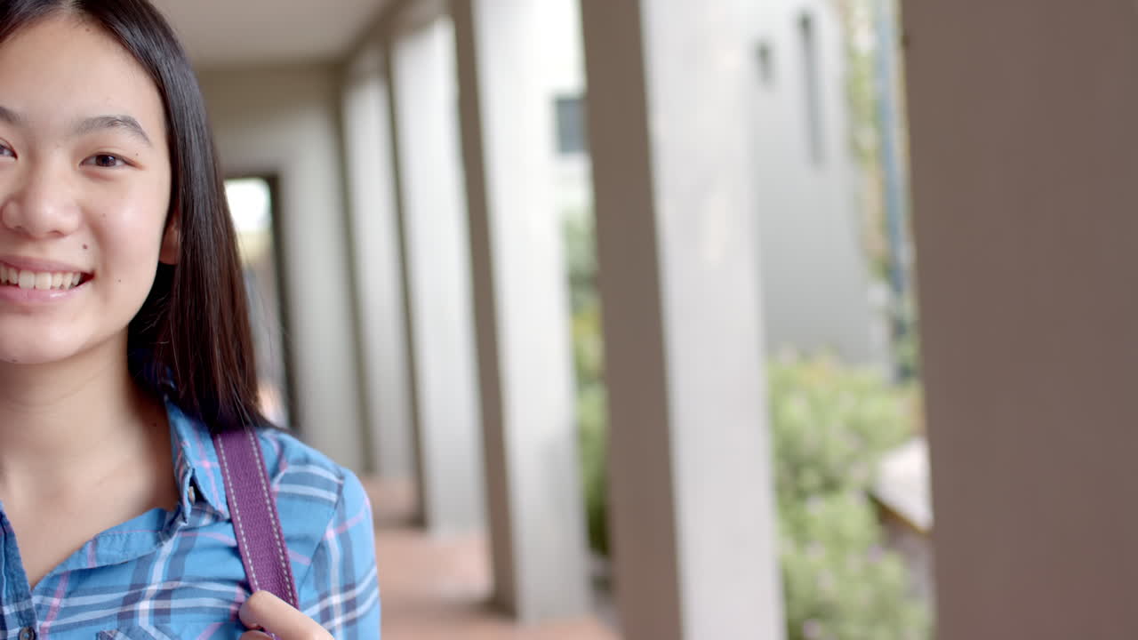 In a highschool corridor, a young female Asian student smiles brightly, copy space