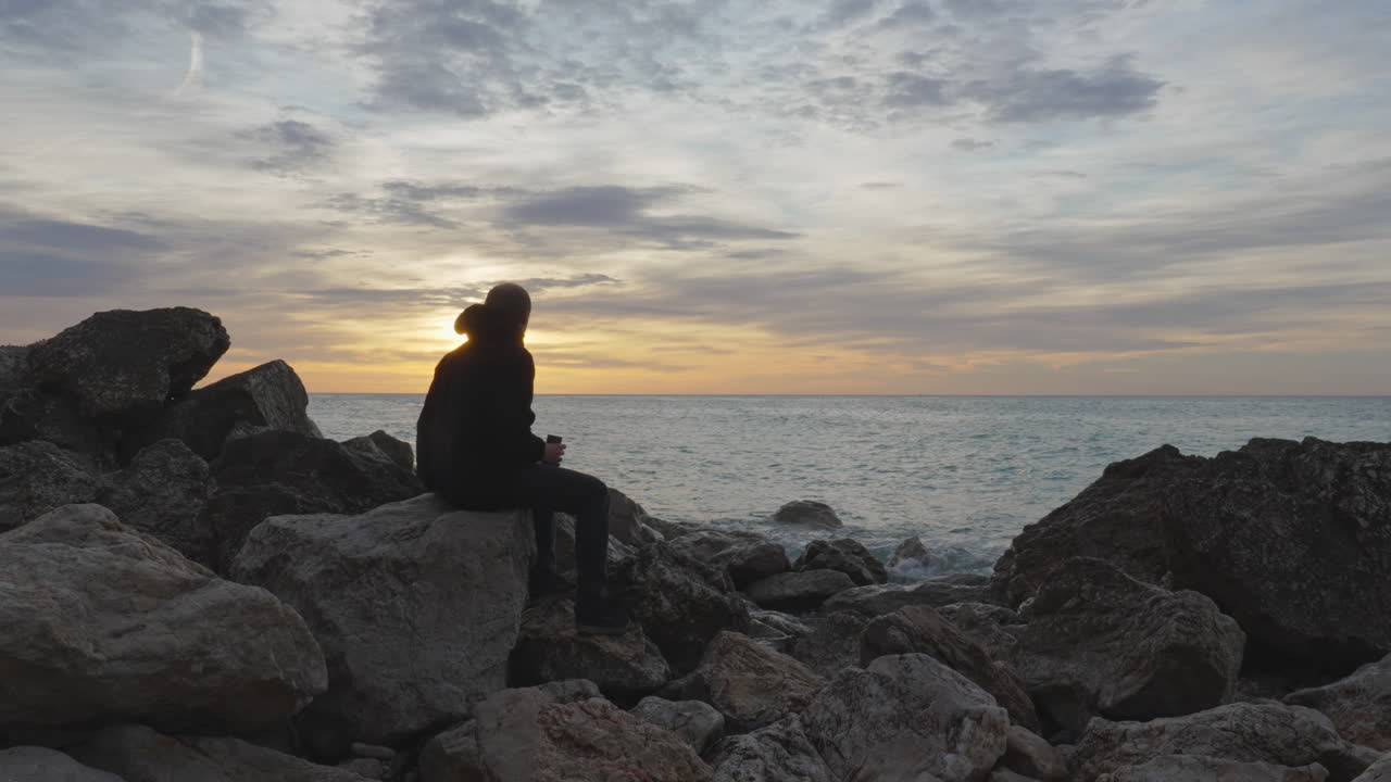 el hombre entra, se sienta en una roca, abre el termo, vierte una bebida caliente y luego la disfruta mientras el mar se lava cerca de sus pies durante la hora dorada