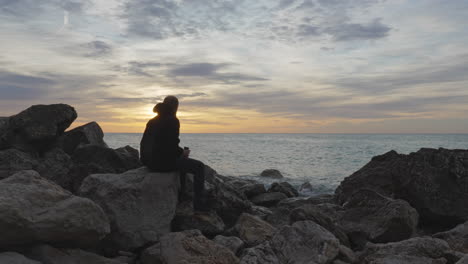 man enters, sits on rock, opens thermos, pours in hot drink, then enjoys it as sea washes near his feet during early during golden hour