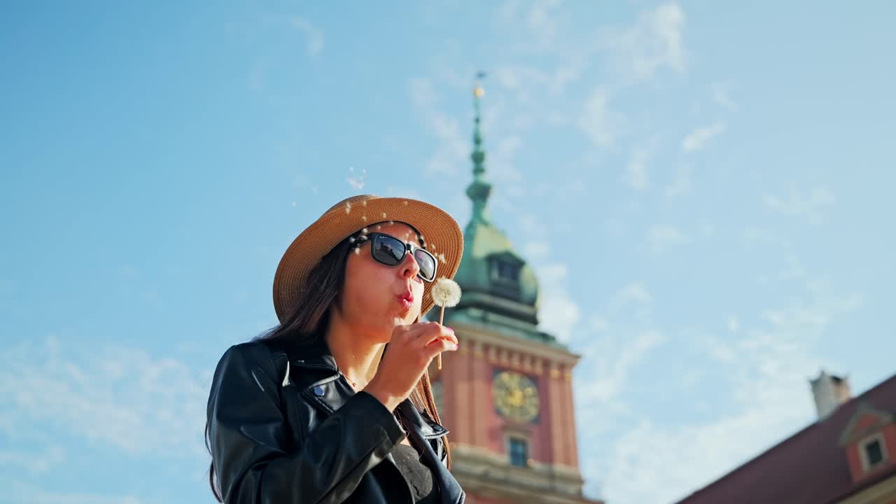 Young woman in hat blowing dandelion near Royal Castle in Warsaw, Poland