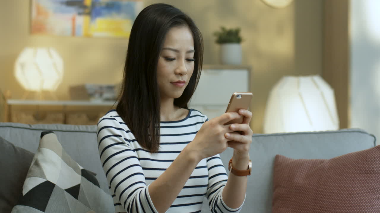 Young Woman In Striped Blouse Tapping And Texting A Message On The Smartphone And Smiling To The Camera Sitting On Sofa In The Living Room
