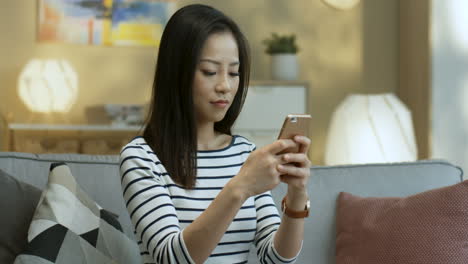 줄무늬 블라우스를 입은 젊은 여성이 스마트폰으로 메시지를 탭하고 문자를 보내고 거실 소파에 앉아 카메라를 보며 웃고 있습니다.