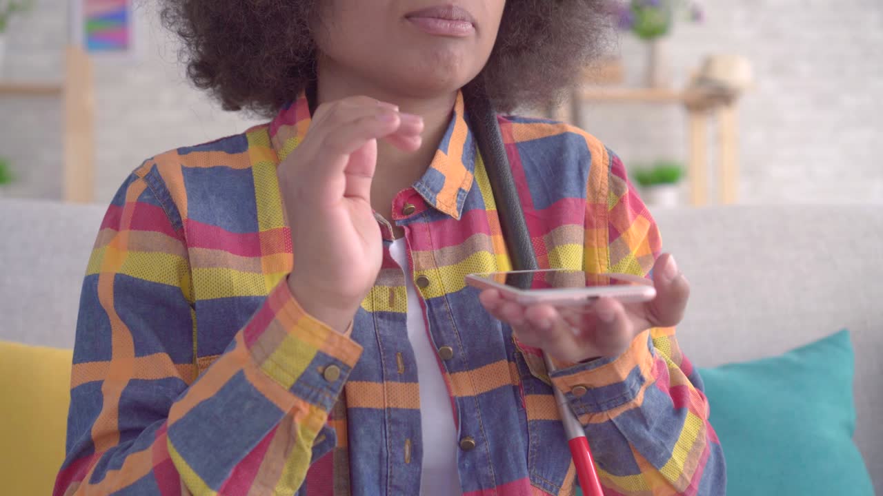 mujer africana ciega con un peinado afro usa asistente de voz en su teléfono inteligente sentada en el sofá de cerca