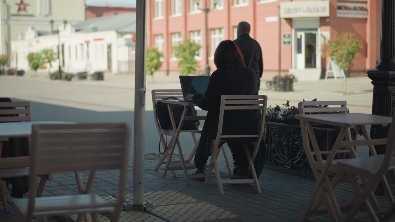 freelancer trabajando al aire libre en una computadora portátil en una cafetería rodeada de sillas de madera, plantas en macetas y paisajes de calles urbanas, con personas caminando en el fondo bajo la luz del sol