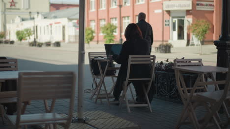 freelancer working outdoors on laptop at cafe surrounded by wooden chairs, potted plants, and urban street scenery, with people walking in the background under sunlight