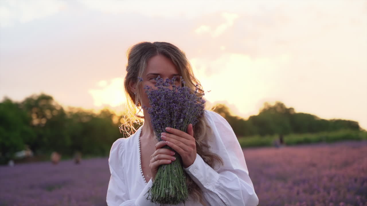 Woman in a white dress holding and smelling a bouquet of lavender in a field at sunset