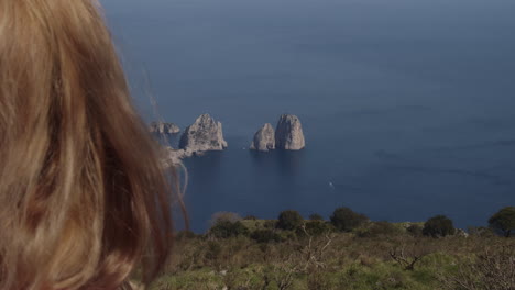 blonde woman sits at mountain overlook, enjoys ocean and rock spire view, capri italy