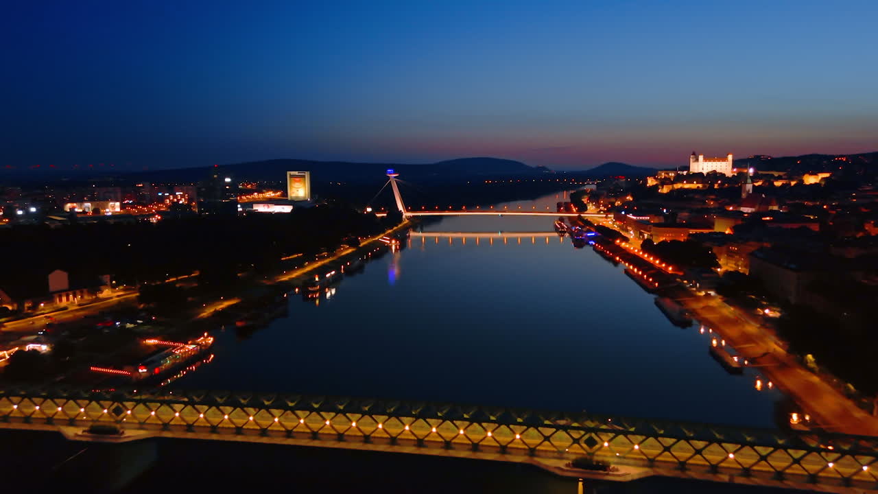 Flight over the calm waterscape of the Danube with the view on the UFO bridge and Old Bridge. Old Castle is well-illuminated on the hill of the historical part of Bratislava, Slovakia
