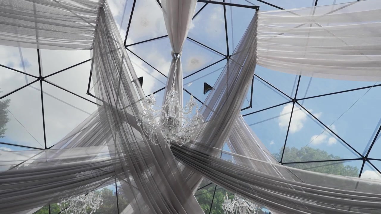 Lampadari di cristallo pendono da un soffitto a cupola di vetro geometrica adornato con drappeggi traslucidi e fluidi, con luce naturale che filtra e alberi rigogliosi visibili all'esterno. Nessuna persona presente.