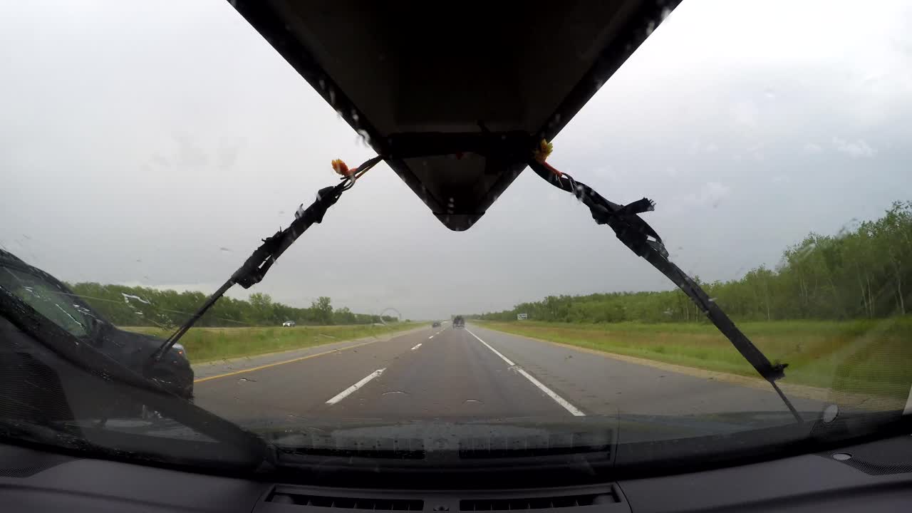 Timelapse GoPro dall'interno del parabrezza di un'auto che percorre la Trans-Canada Highway attraverso le praterie canadesi. Canoa legata al tetto, terreni agricoli pianeggianti, alberi a bordo strada, pioggia battente e traffico sotto un cielo coperto.