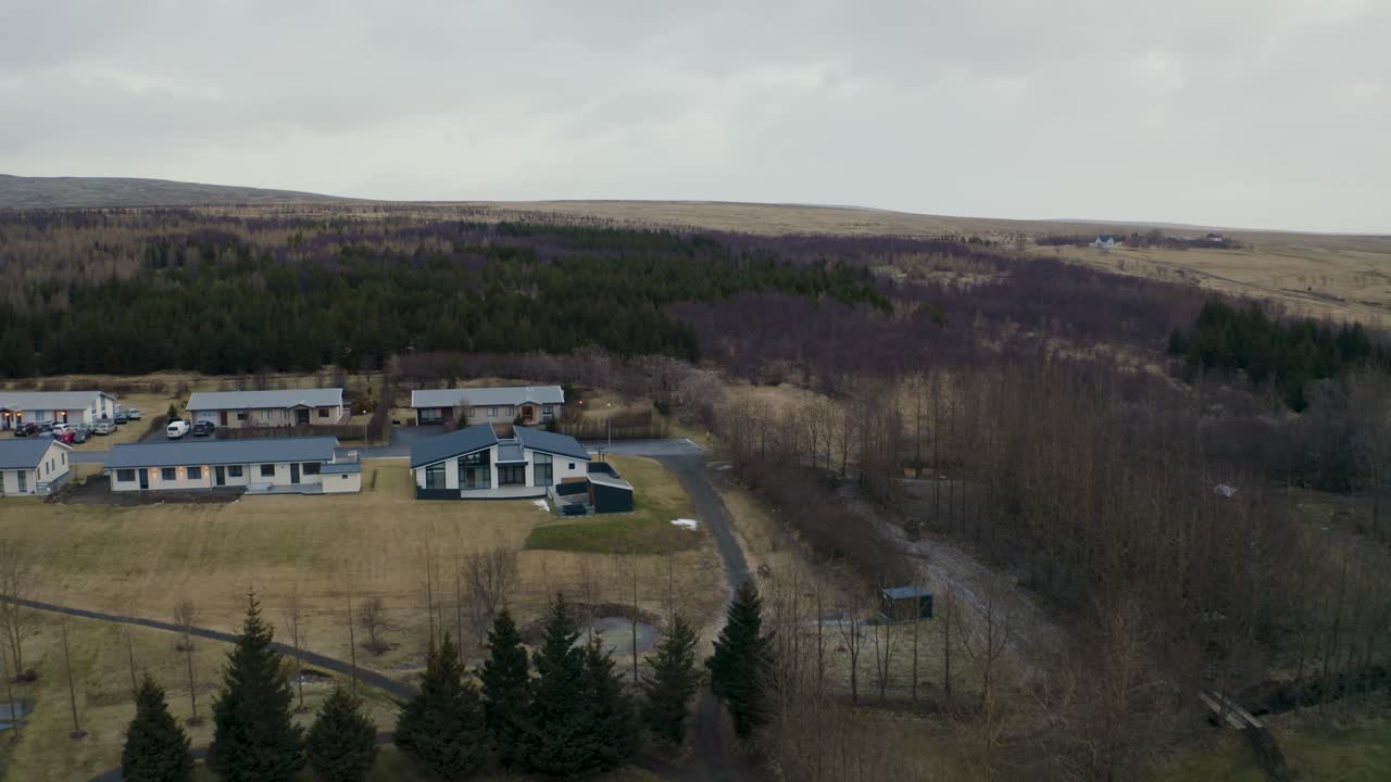 Drone footage showcasing a peaceful rural village in Iceland, surrounded by dense forests, open fields, and scattered homes. A serene landscape blending nature and human settlement.
