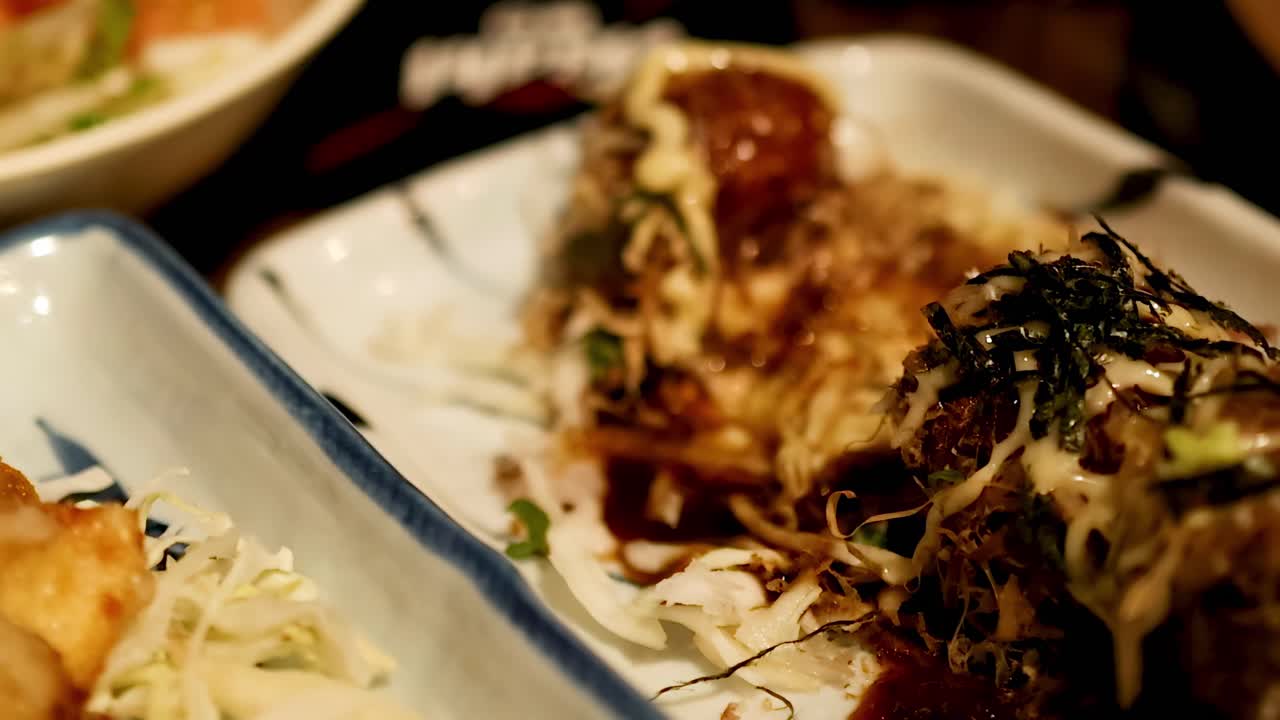Close-up of chopsticks picking up takoyaki, topped with sauce and seaweed, from a garnished plate.