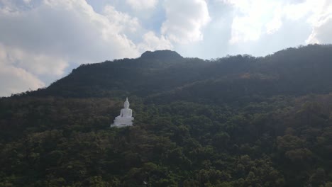 Luang-Por-Khao,-Wat-Theppitak-Punnaram,-sliding-aerial-footage-to-the-left-of-the-famous-white-Buddhist-shrine-on-the-mountain-side-revealing-an-afternoon-summer-sky-and-lovely-changing-colour-trees