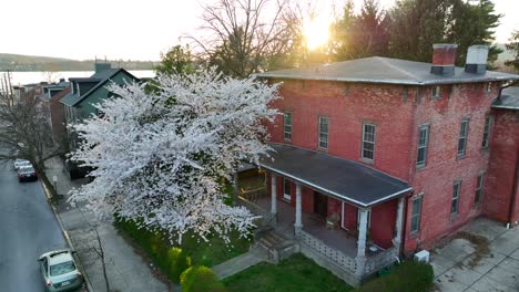 drone view rising up of a historic red brick house with a large blossoming tree in the front yard and the surrounding neighborhood and reflections of a river during sunset