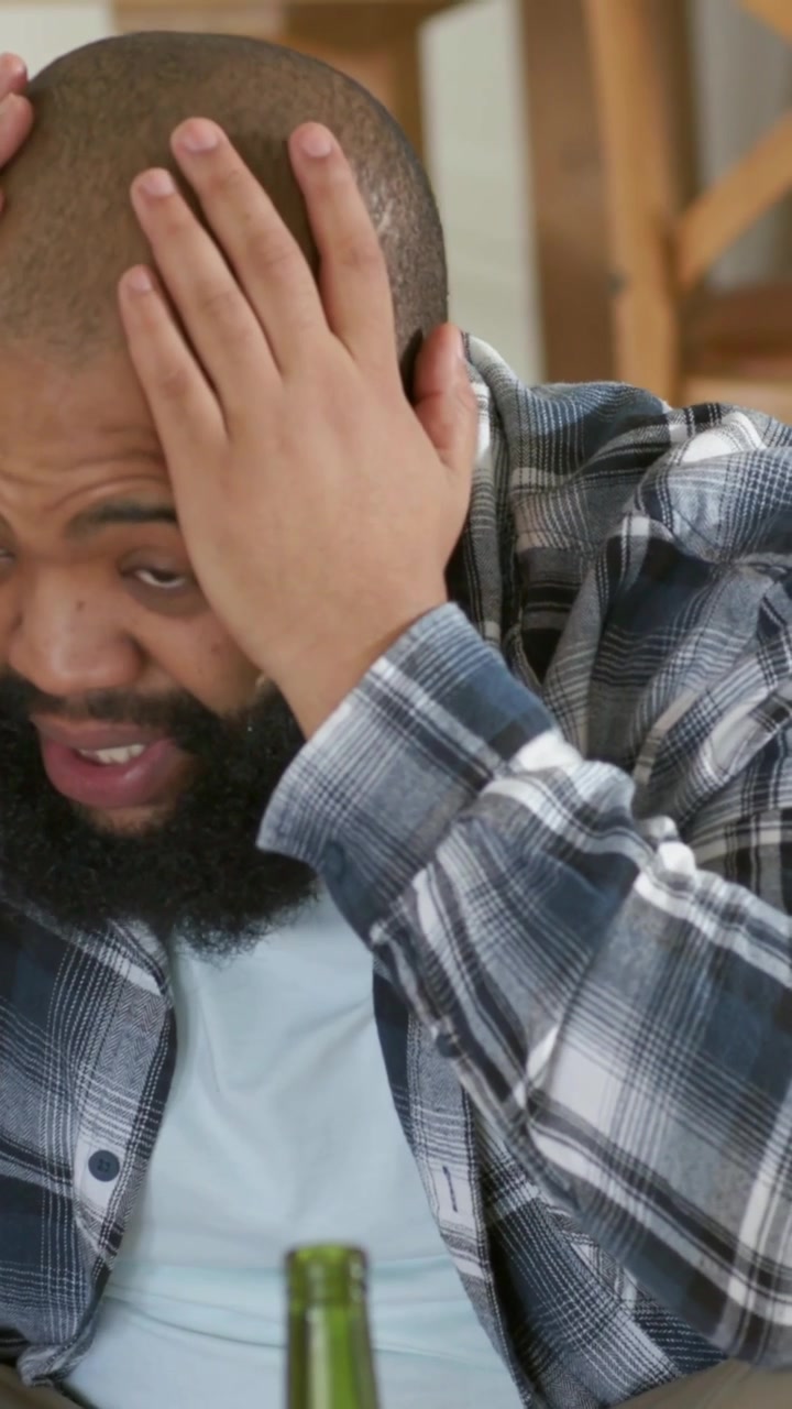 Vertical video: Raising right hand, man in plaid shirt covering face on tan couch with green bottle