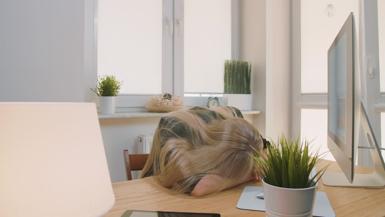 Tired woman waking up at workplace. Tired blond female office worker in elegant suit relaxing lying on arms on desk then getting up yawning and starting working on computer in light room with plants.