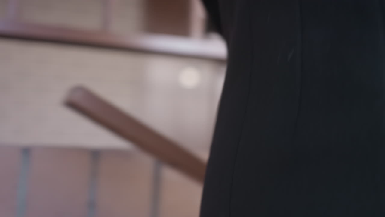 Mujer serena con abrigo negro abre un bolso bandolera de cuero marrón en un pasillo de ladrillo, con manos cuidadosas buscando dentro del bolsillo con solapa; momento cotidiano, primer plano medio centrado en el gesto y los detalles del accesorio