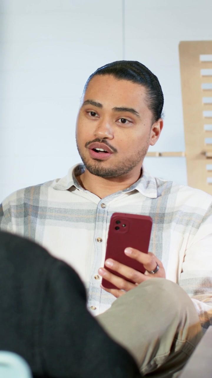 Video verticale: Uomo con camicia a quadri che guarda in alto e parla in un salotto, tenendo un telefono rosso per un segnale