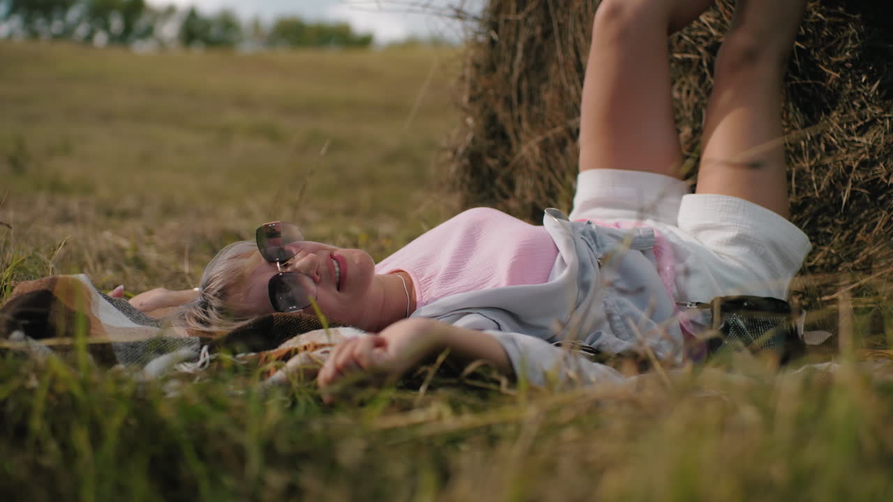 женщина, лежащая на коврике в травяном поле, в солнцезащитных очках, расслабленная с одной рукой, касающейся травы, ноги, опирающиеся на сено, наслаждающаяся спокойной сельской атмосферой под теплым солнечным светом