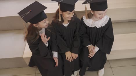 three happy little girls in cap and gown sitting on stairs and talking together at the preschool graduation ceremony