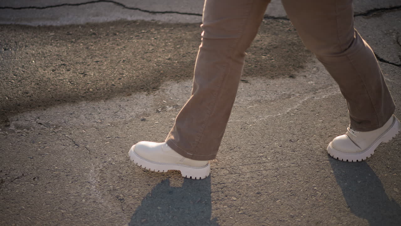 Cracked pavement under white boots as female dancer expresses rhythmic motion through graceful footwork on sunlit urban street, winter pants swaying, shadows stretching across melting snow lined curb