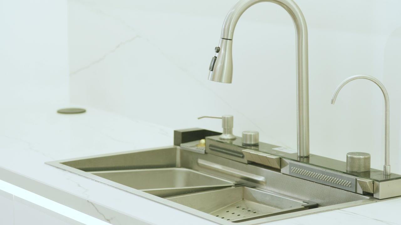 Encuadre detallado que muestra un área de lavado con doble fregadero de acero inoxidable, grifo alto y flexible, y controles auxiliares, sobre una encimera blanca de líneas limpias