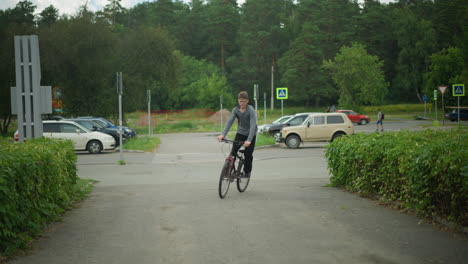 young boy wearing glasses rides bicycle on interlock path, surrounded by greenery, parked cars, pedestrians, signpost in background, quiet suburban street, with greenery in the background