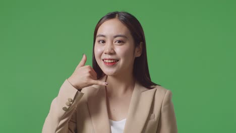 아시아 사업가 여성이 따뜻하게 미소 짓고 스튜디오에서 녹색 화면 배경에 전화 제스처를 하고 있습니다.