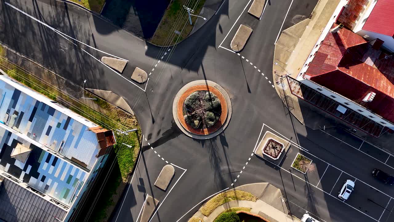 Overhead drone footage captures vehicles navigating a small city roundabout surrounded by commercial buildings in bright daylight, highlighting urban infrastructure and traffic flow