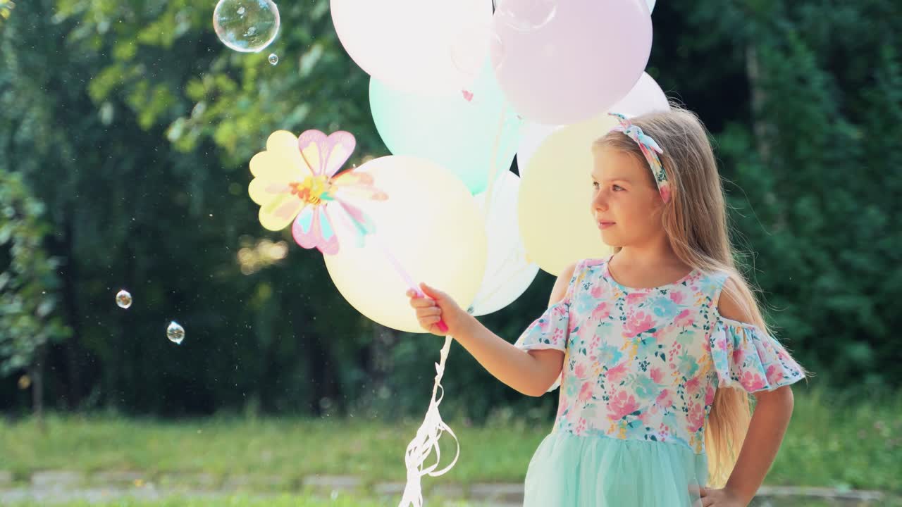 Little girl with balloons in her hands blows a multi-colored toy windmill in nature against the backdrop of trees. Flying soap bubbles