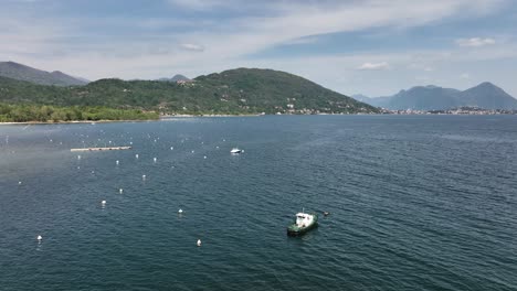 Low-aerial-orbit-of-a-boat-anchored-on-a-giant-mountain-lake-with-buoys-tree-covered-hills,-distant-mountains-and-villages