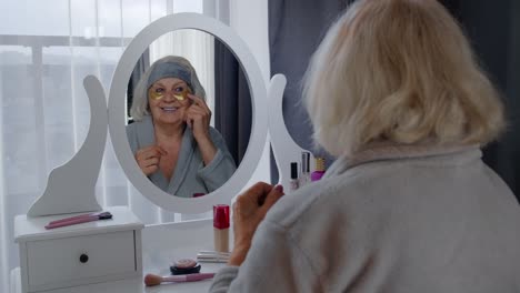 an elderly woman applies gold under eye patches in her bathroom