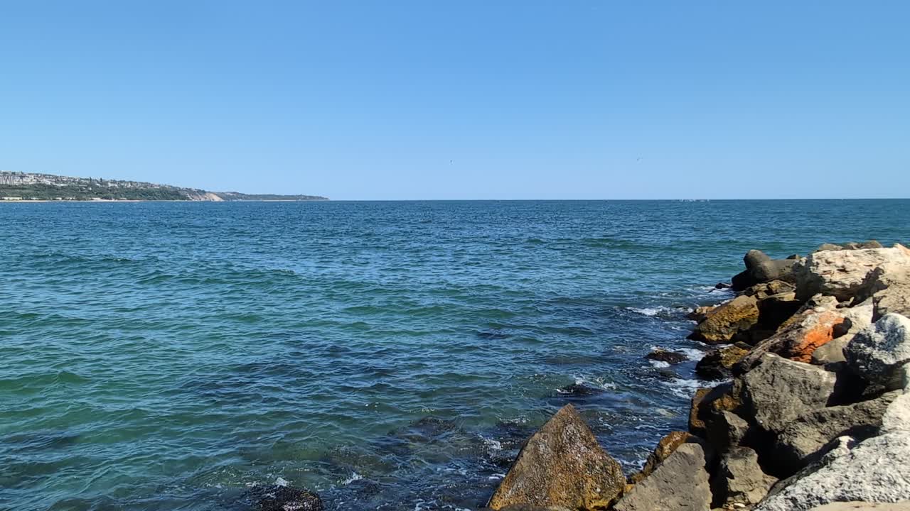 Kayalık deniz duvarı ve berrak mavi suyu ile Karadeniz'in kıyıdan görünümü. Bulgaristan kıyısı boyunca sakin, doğal bir deniz manzarası yaratan hafif dalgalar taşlık sahille buluşuyor