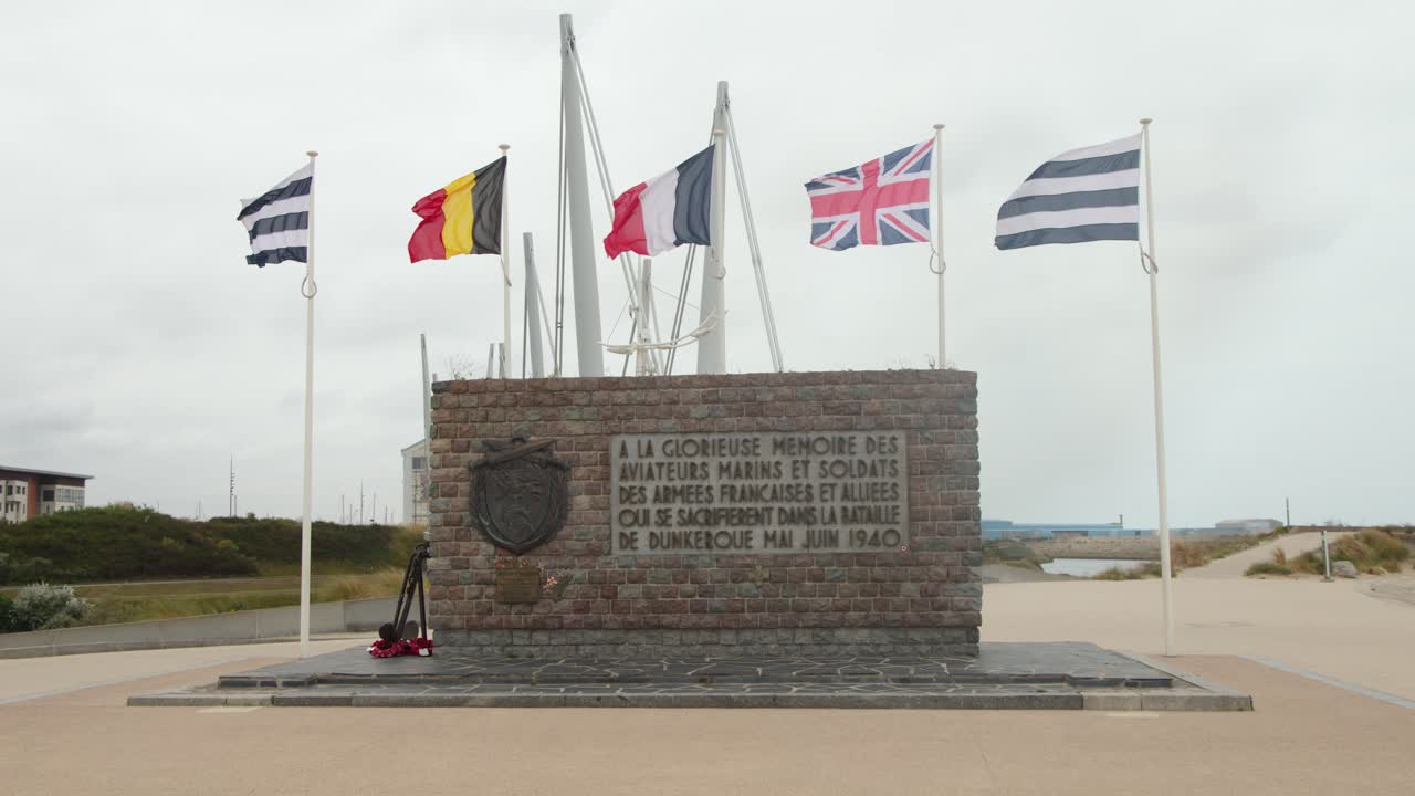 Static wide shot of coastal war memorial with multiple flags waving, overcast daylight, no people