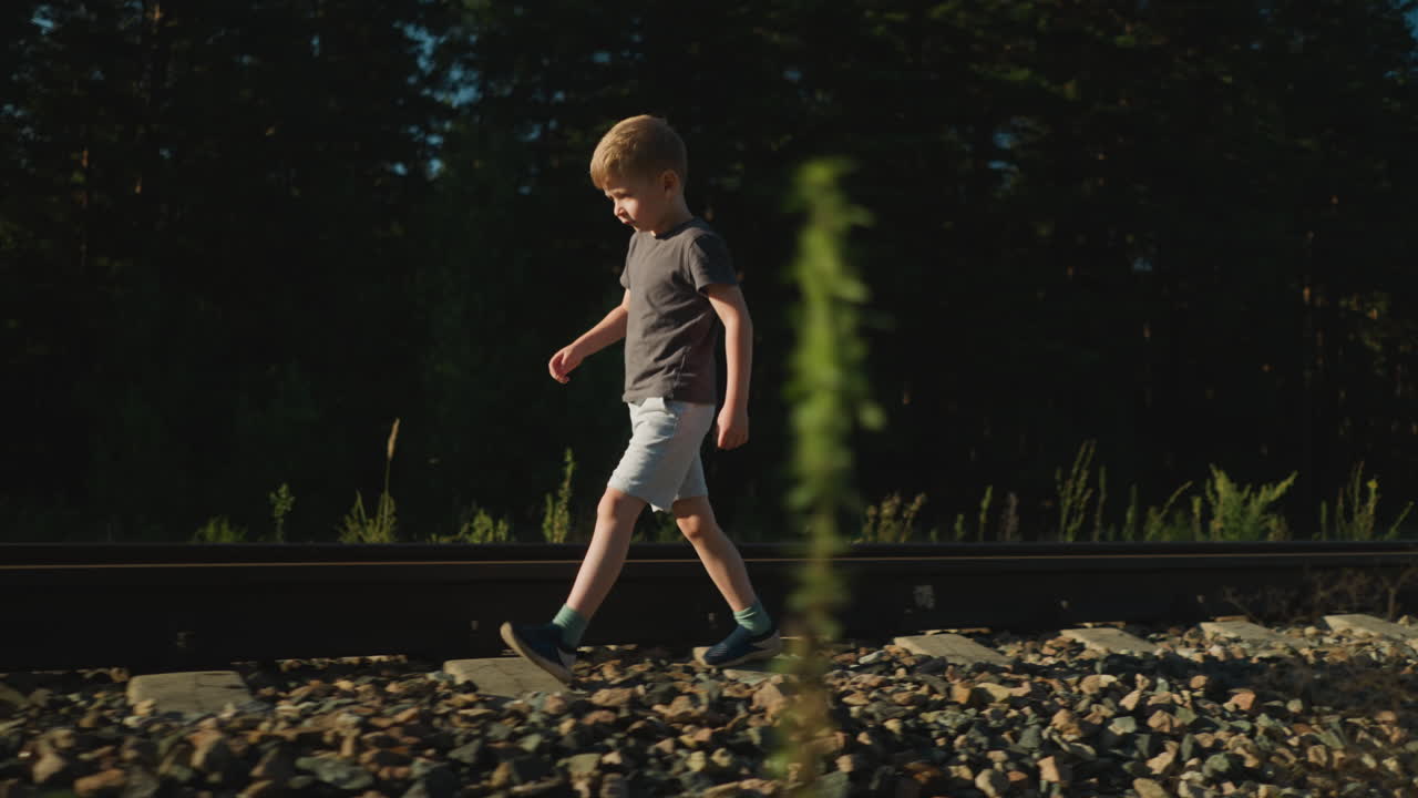 Side view of young boy in casual outfit walking beside railway track on rocky path surrounded by wild vegetation and forest trees during sunny day with soft shadows and natural lighting
