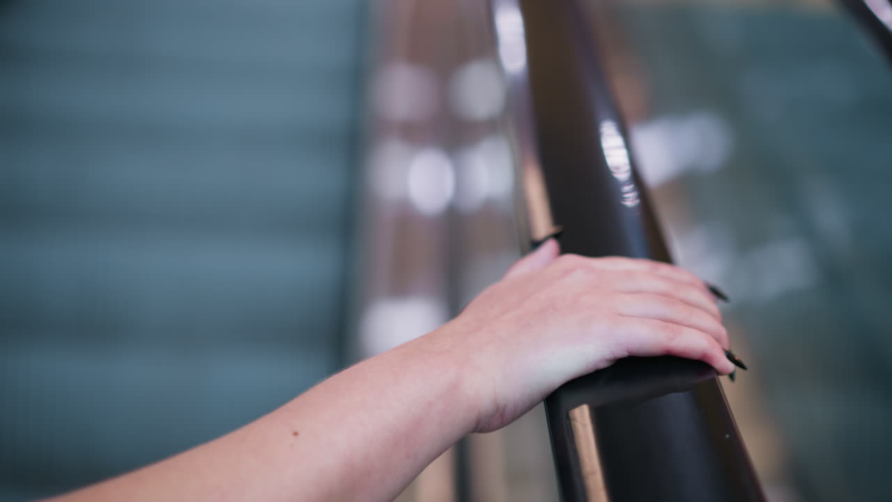 primer plano de la mano derecha de la dama descansando en el riel de la escalera mecánica con uñas pulidas negras, textura suave de la piel y venas sutiles visibles, fondo con luces bokeh y reflejos metálicos