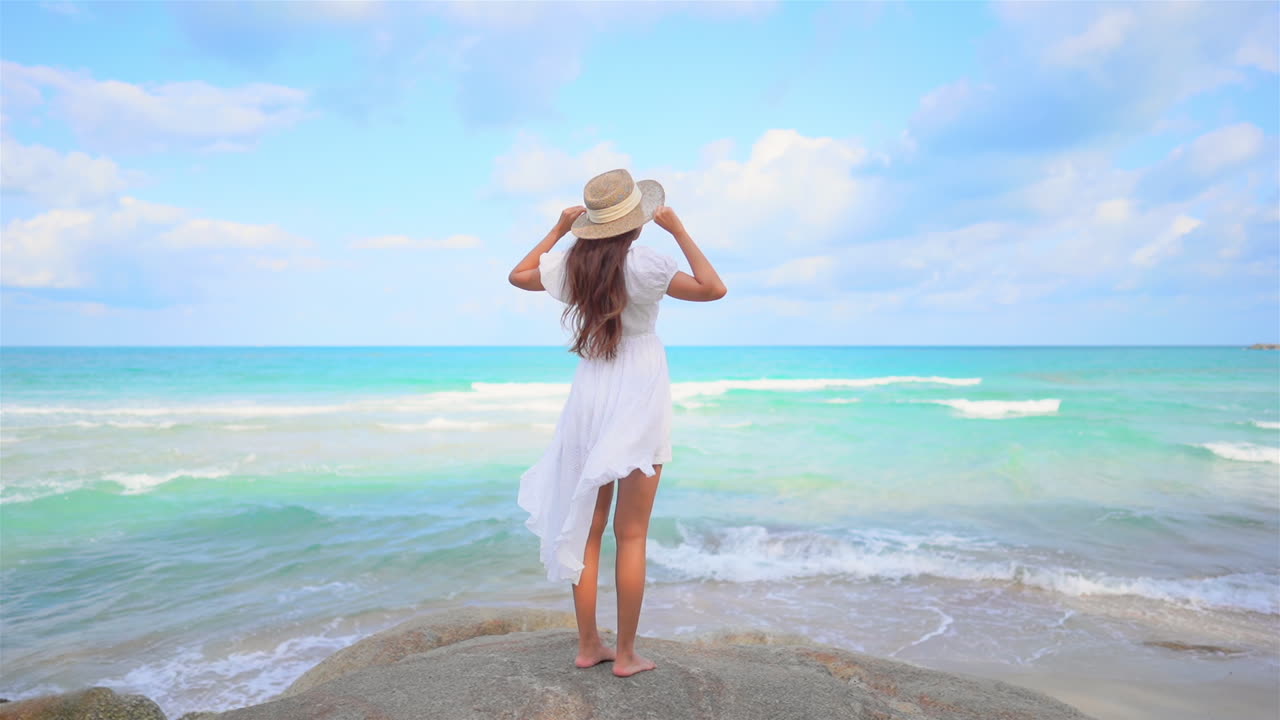 하얀 선드레스와 짚으로 된 태양 모자를 쓴 젊은 여성이 다가오는 파도가 내려다보이는 바위 위에 서 있습니다.