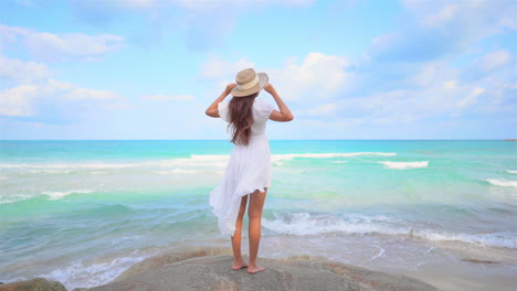 A-young-woman-in-a-white-sundress-and-straw-sun-hat-stands-on-a-boulder-overlooking-the-incoming-surf