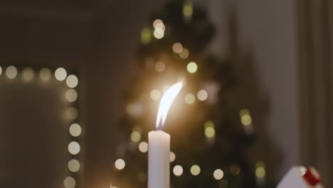 The-Camera-Focuses-On-A-Burning-Candle,-There-Is-A-Christmas-Tree-With-Blurred-Lights-In-The-Background