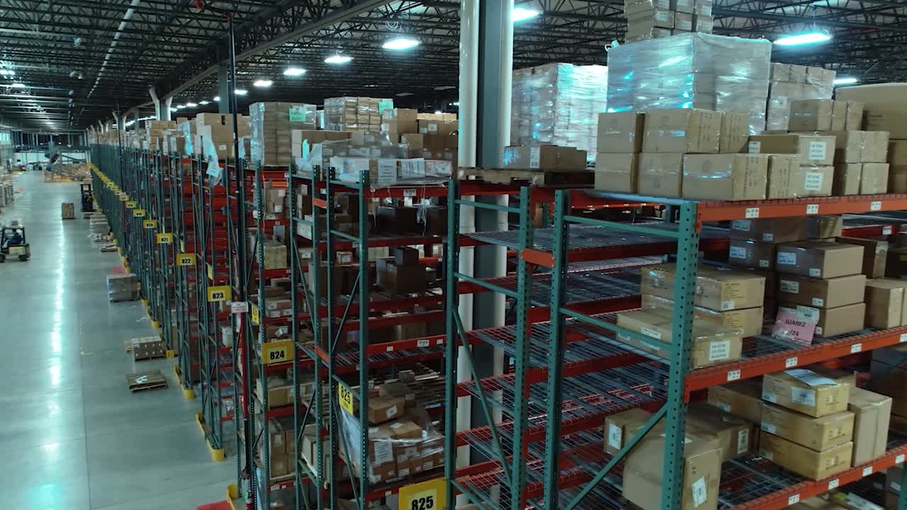 A rotating drone shot around the end of a tall warehouse shelving row, revealing multiple aisles, moving forklifts, and stacks of organized packages