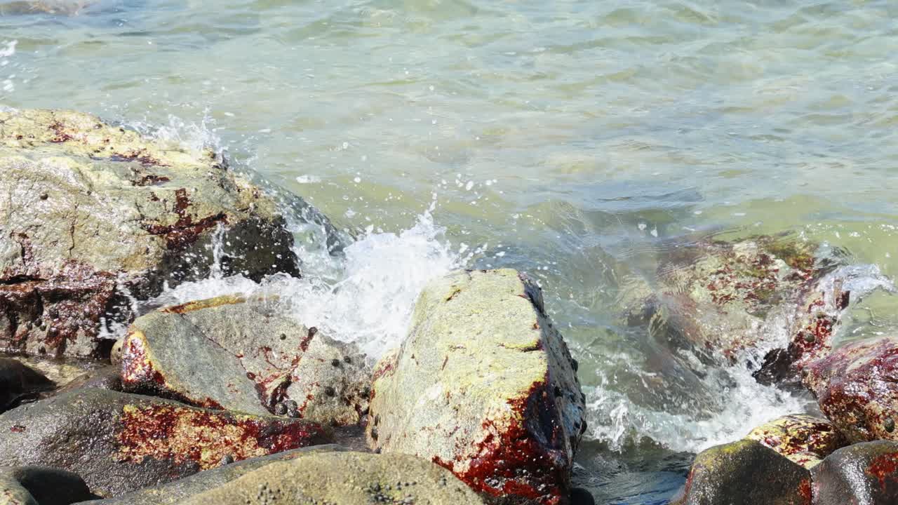 Waves crash against algae-covered rocks on a sunny day in Phuket, Thailand, creating a dynamic and refreshing scene