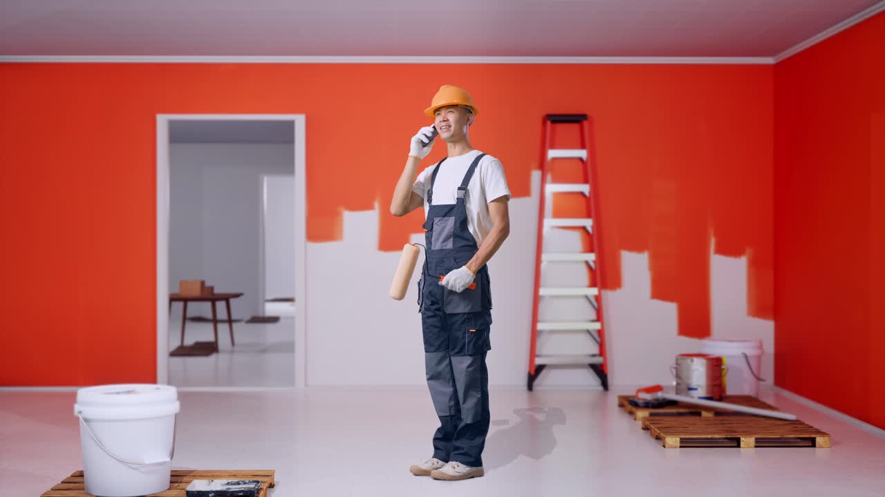 Full Body Of Side View Asian Man Painter Wearing Safety Helmet Talking On Smartphone And Looking Around While Standing With Painting Wall In Room