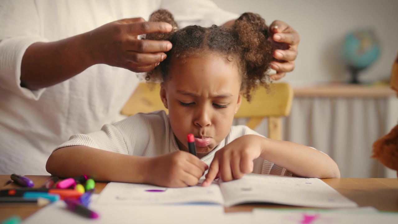 Mãe penteando o cabelo da filha enquanto ela desenha em casa