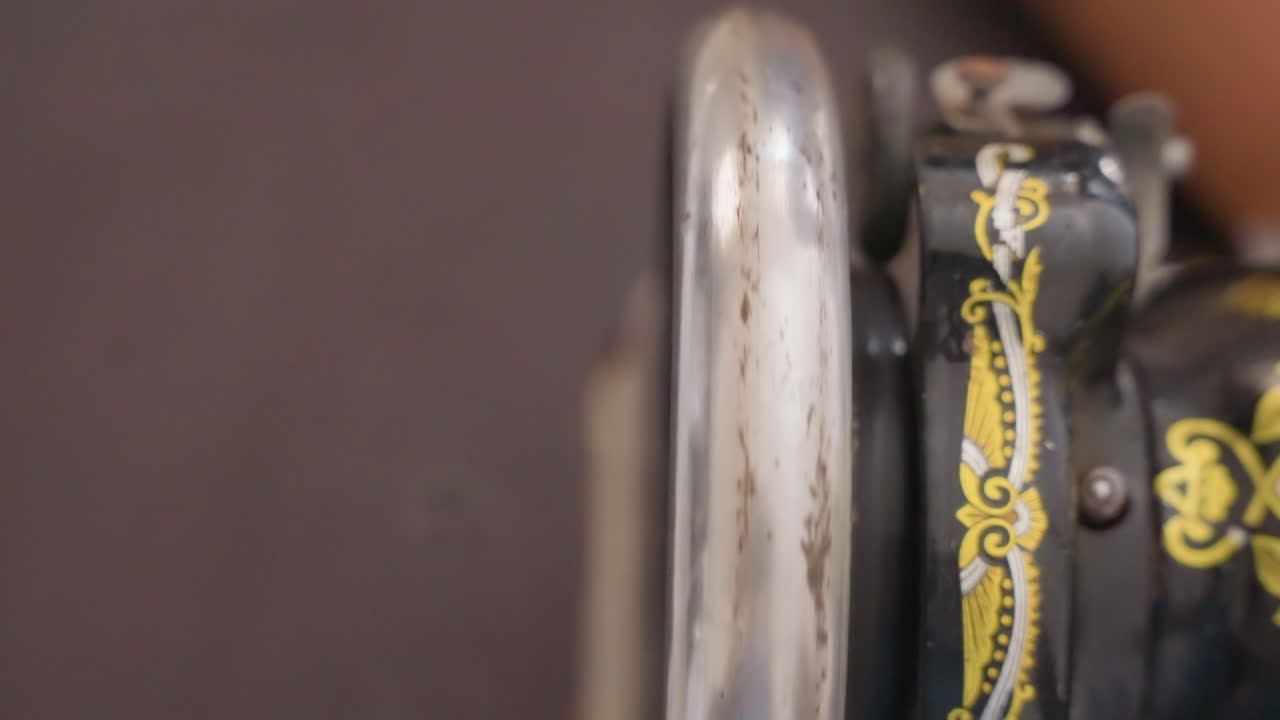 Closeup black sewing machine wheel showing worn chrome and ornate yellow decal, slow motion rotation, domestic maintenance scene in quiet home workshop, muted background, tactile metal patina