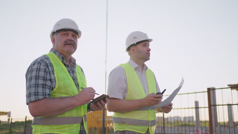 Retrato-De-Dos-Constructores-De-Pie-En-El-Sitio-De-Construcción.-Dos-Constructores-Con-Dibujos-Parados-En-El-Fondo-De-Edificios-En-Construcción-Con-Cascos-Y-Chalecos