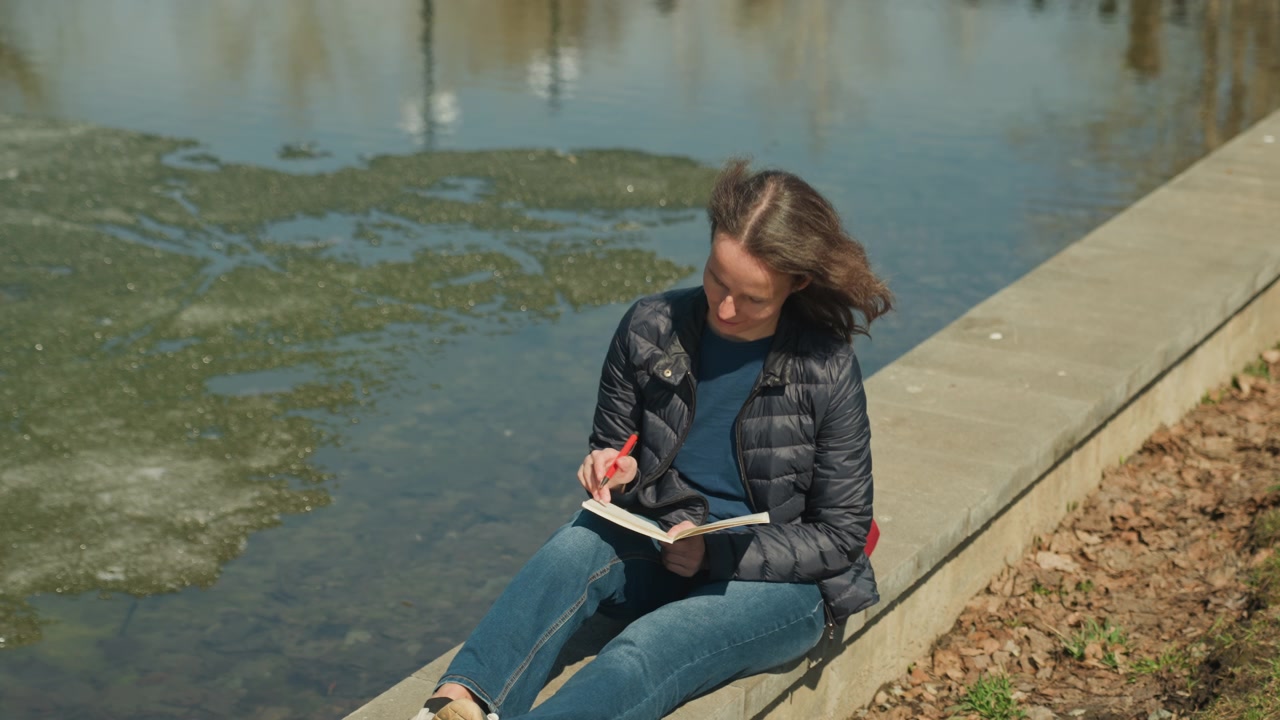 Primo piano di una donna seduta vicino all'acqua con un taccuino e una penna, che osserva l'ambiente circostante. La donna tiene un libro aperto in grembo mentre impugna uno strumento di scrittura rosso; i lunghi capelli si muovono leggermente, indicando una brezza.
