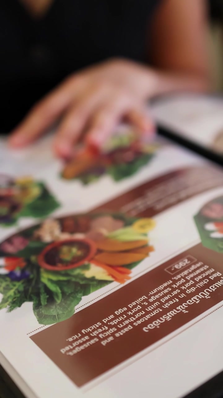 En person bladrer igennem en farverig nordthailandsk madmenu ved et restaurantbord, med naturligt lys og lav dybdeskarphed, der fremhæver de livlige retter.