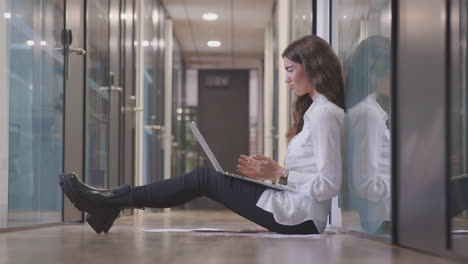 Frustrated-Young-Businesswoman-Sitting-On-Floor-In-Corridor-Of-Modern-Office-Working-On-Laptop