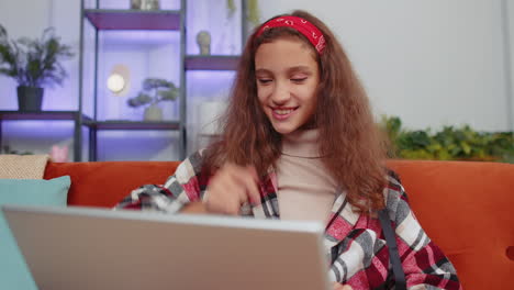 mujer joven sentada en el sofá, sonriendo, mirando una computadora portátil, saludando, pensando, teniendo una llamada de video en línea