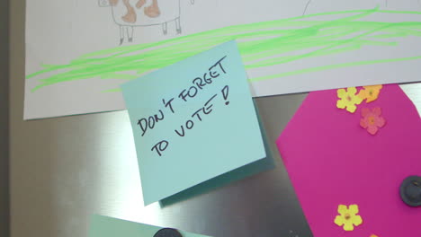 a person's hand sticks a reminder on the refrigerator door with the handwritten message, don't forget to vote